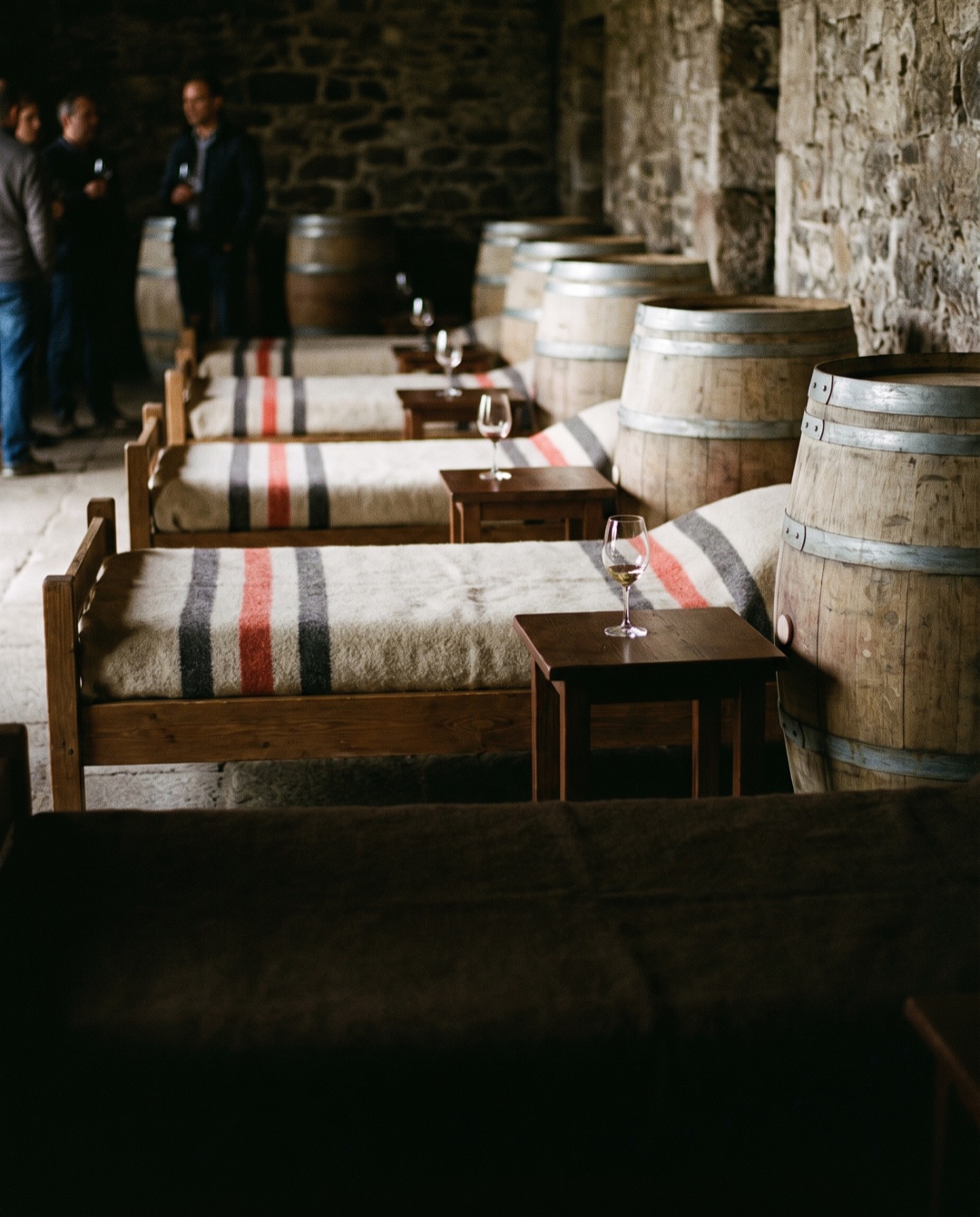 Cushioned daybeds arranged between oak barrels in a dim cellar, each with a folded blanket and a single wine glass.