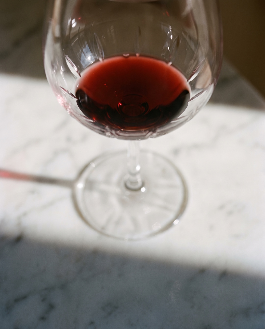 A single ounce of red wine barely covers the bottom of a large crystal stemmed glass on white marble.