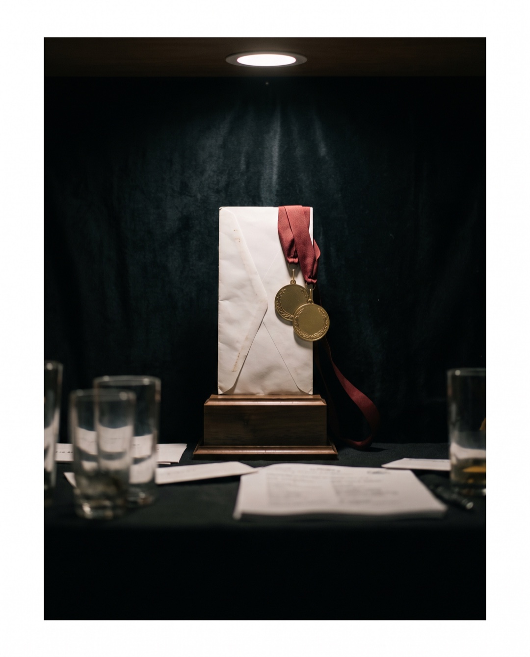 A white business envelope stands upright on a wooden trophy plinth with two gold medals on ribbons draped over its corner.