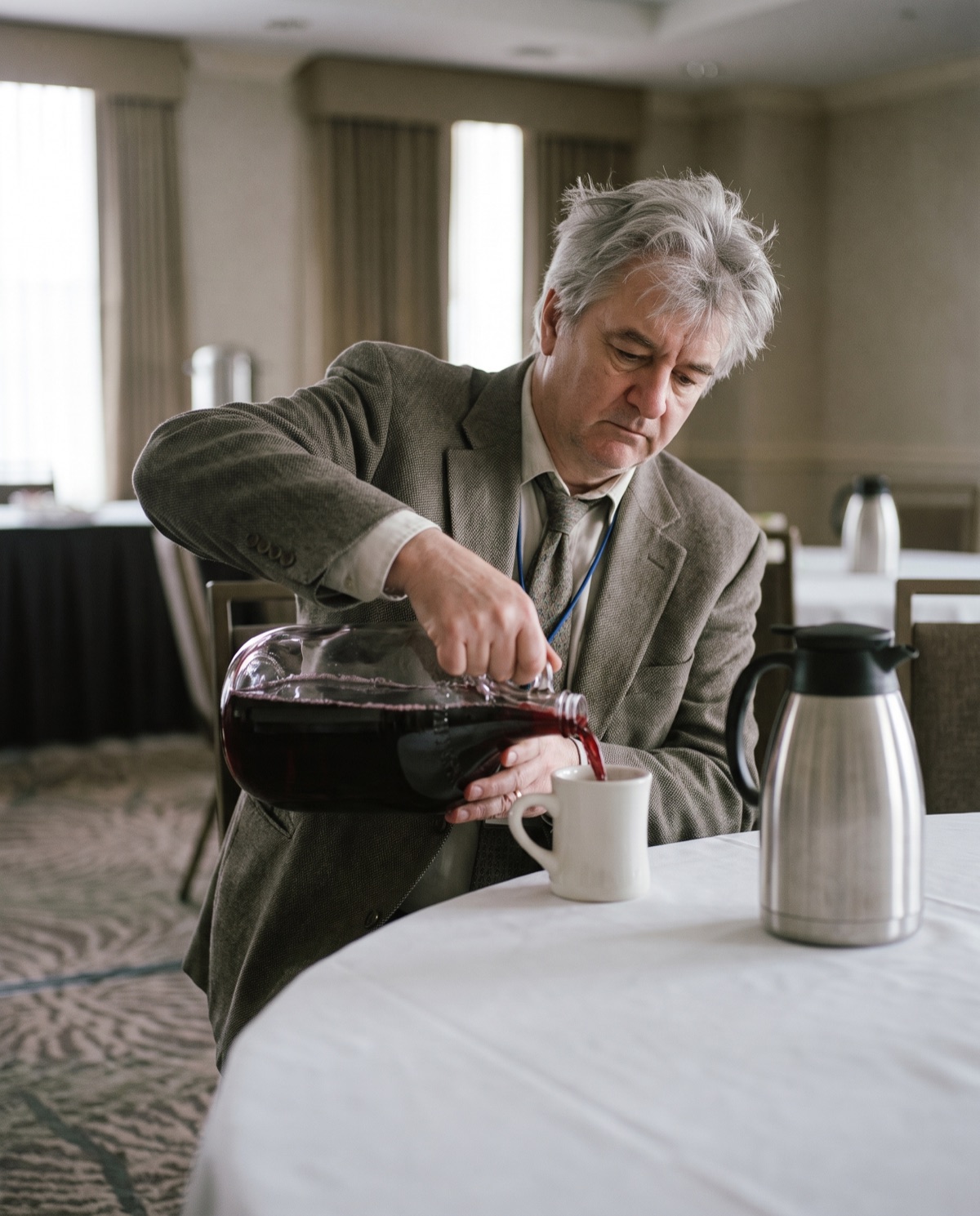 A man pours red wine from a large glass jug into a white coffee mug beside a stainless coffee carafe.