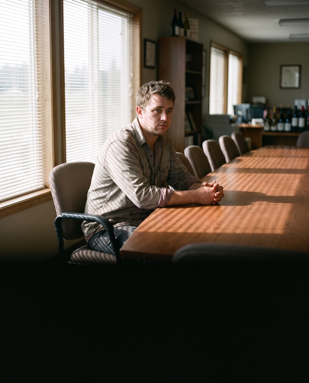 A young cellar worker sits alone at the head of a long empty conference table with ten empty chairs.