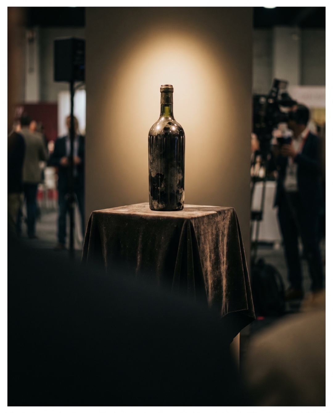 A single heavy thick-glassed wine bottle on dark velvet under a warm spotlight on a display pedestal.